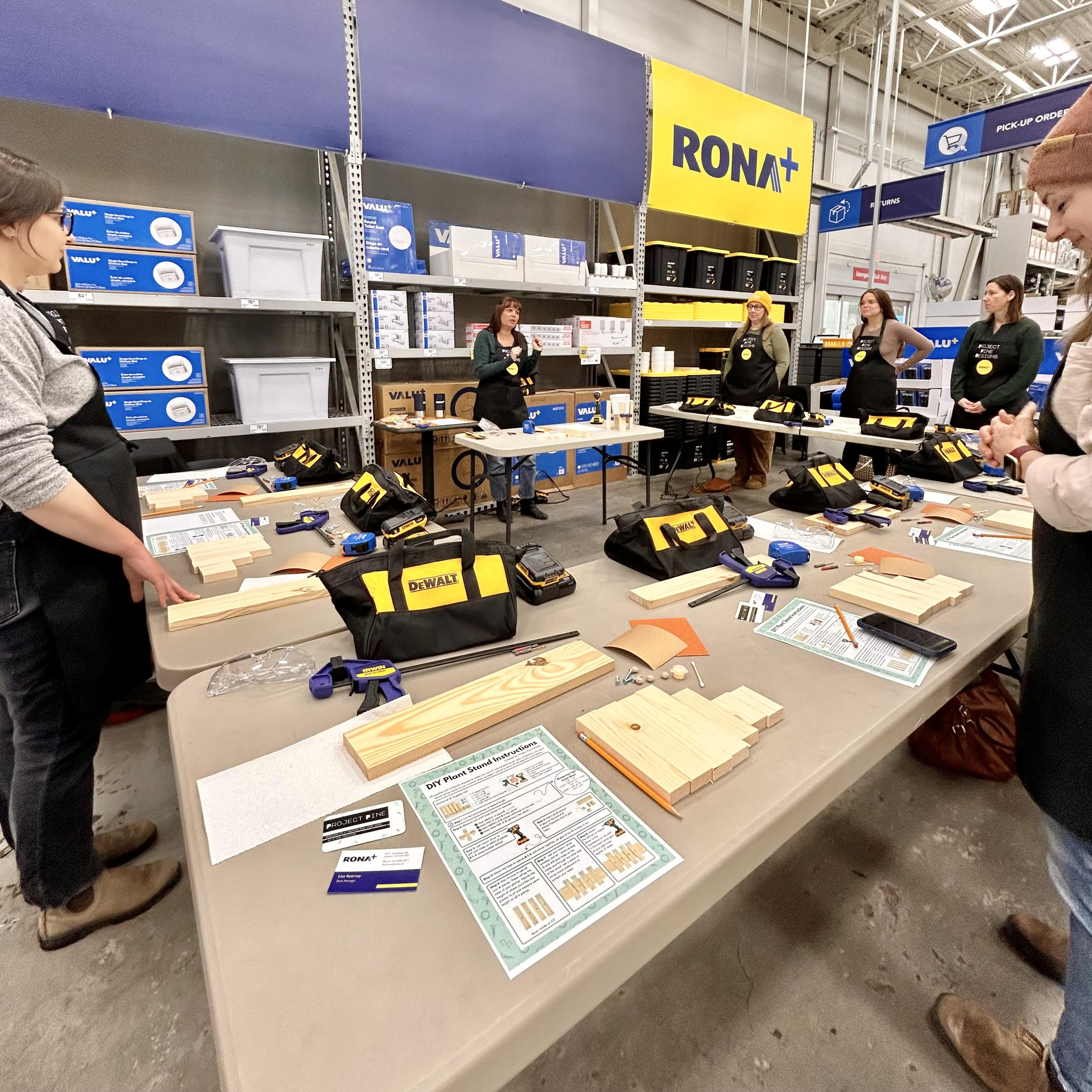 Group of women standing at tables in a U shape with materials to create plant stands watching a female instructor